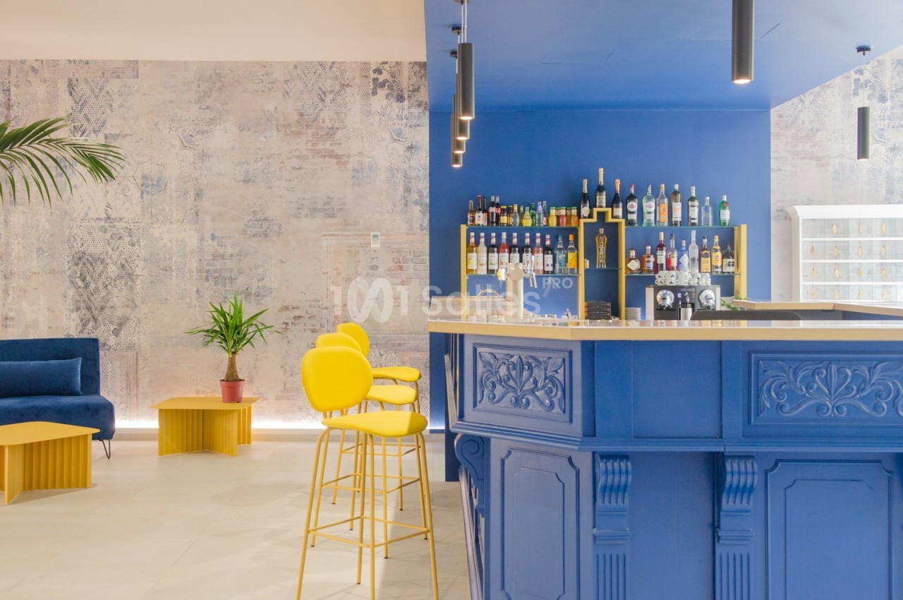 Bar moderne avec comptoir bleu, tabourets jaunes, étagères de bouteilles et mur texturé en arrière-plan.