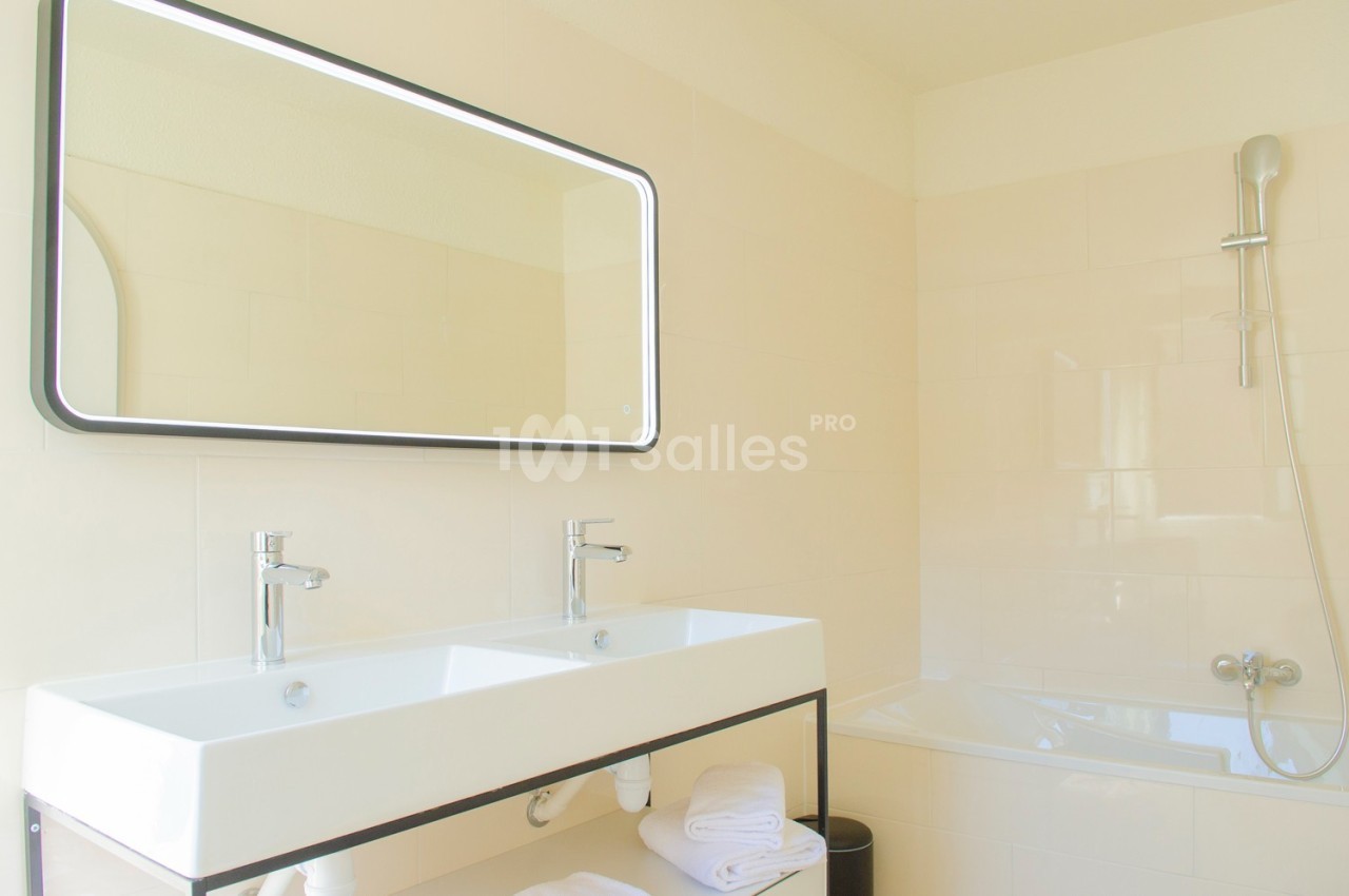 Salle de bain lumineuse avec un double lavabo, un grand miroir, une baignoire et des murs carrelés clairs.