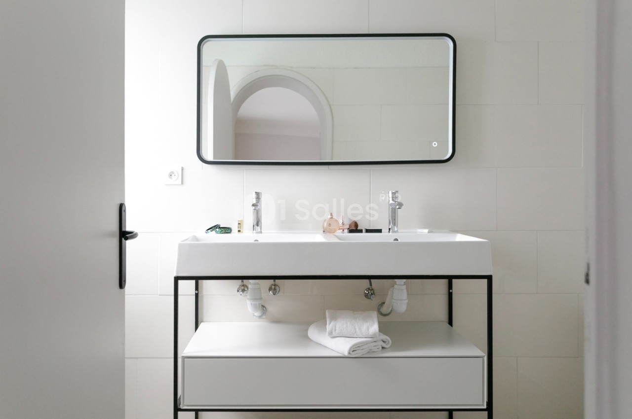 Lavabo double avec miroir rectangulaire, serviette pliée sur une étagère et mur carrelé blanc en arrière-plan.