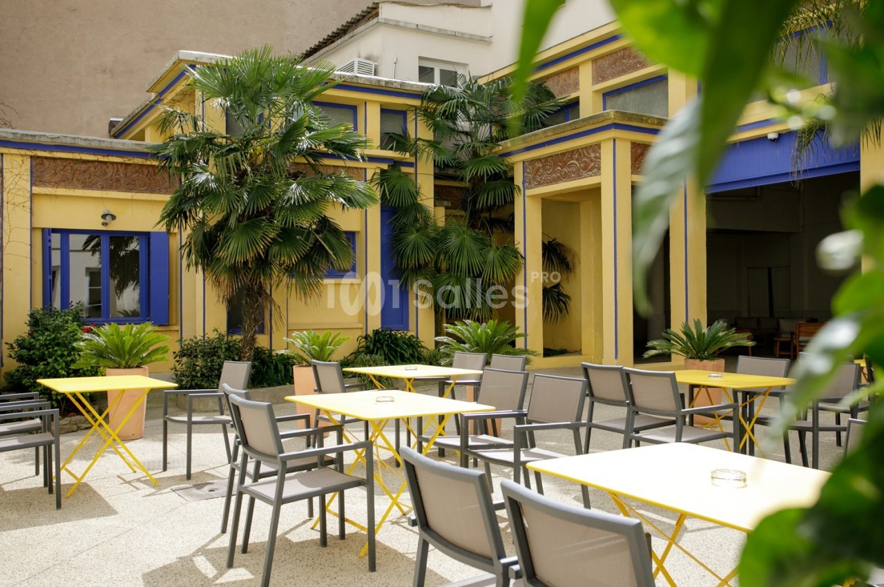 Cour intérieure lumineuse avec tables et chaises jaunes et grises, entourée de plantes et d'un bâtiment coloré.