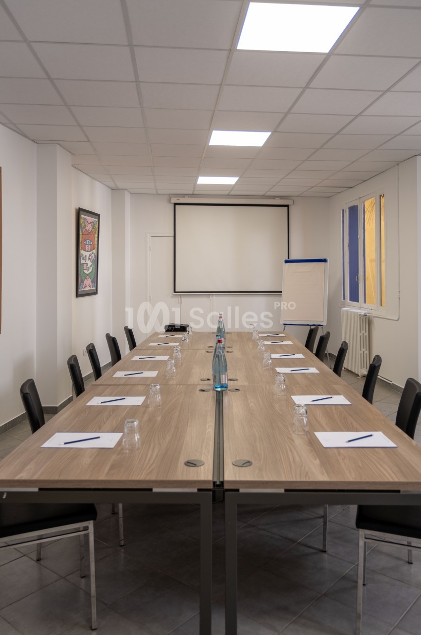 Salle de réunion avec une grande table rectangulaire, des chaises, un tableau blanc et un écran de projection.