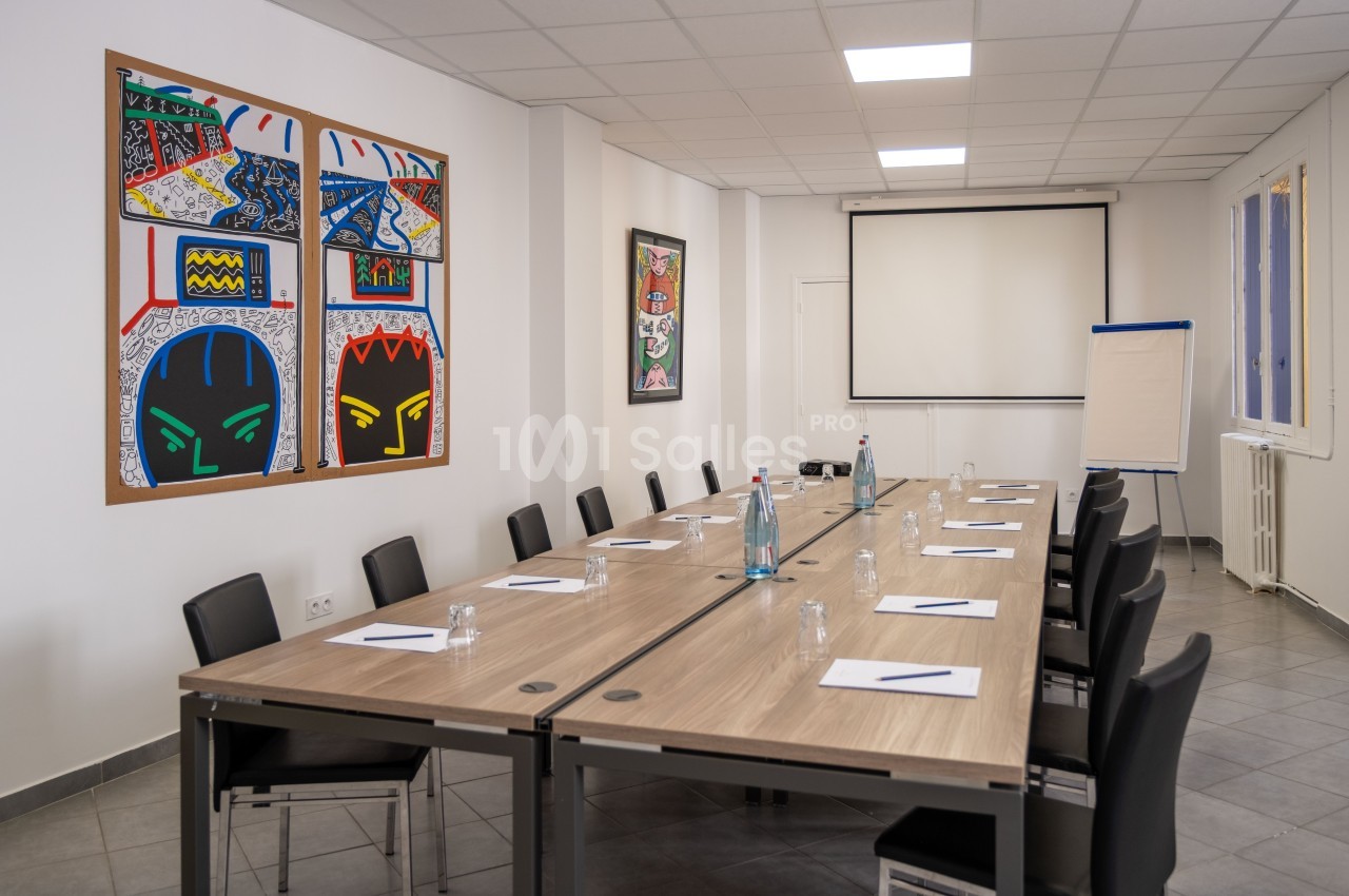 Salle de réunion lumineuse avec une grande table, des chaises noires, un tableau blanc et des œuvres d'art colorées au mur.