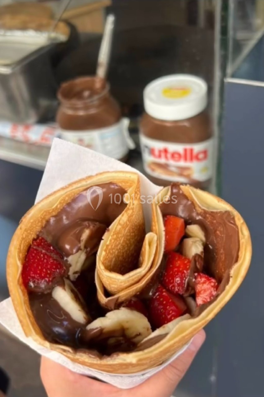 Crêpe garnie de chocolat, fraises et bananes, tenue à la main devant un stand avec un pot de Nutella visible.