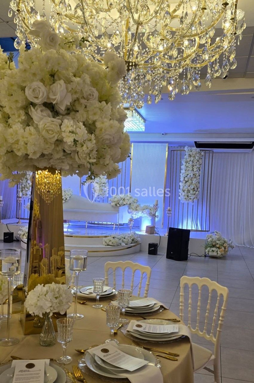 Salle de réception élégante avec décoration florale blanche, lustres en cristal et table dressée avec vaisselle raffinée.