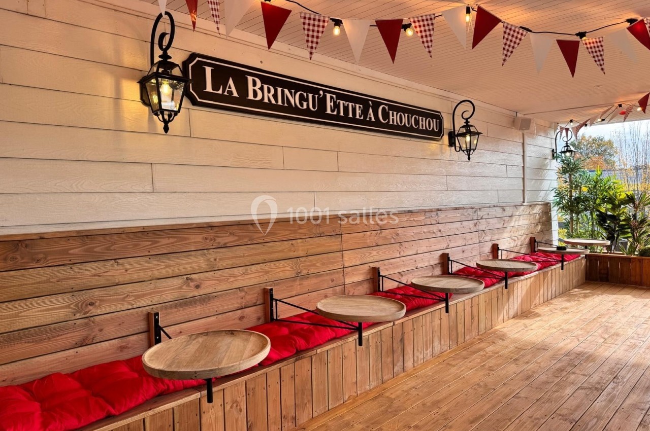 Bancs en bois avec coussins rouges et petites tables rondes, sous une terrasse décorée de fanions et lanternes.