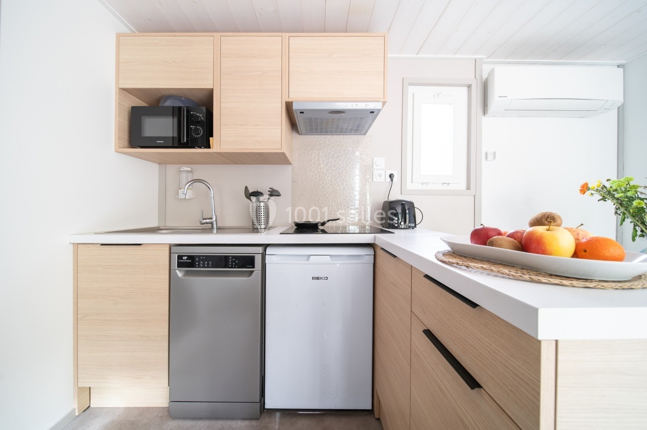 Cuisine moderne avec meubles en bois clair, évier, lave-vaisselle, micro-ondes, réfrigérateur et corbeille de fruits.