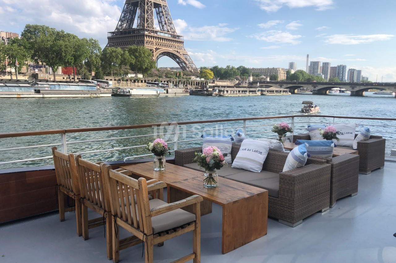 Terrasse d'un bateau avec mobilier en bois et vue sur la Tour Eiffel et la Seine par une journée ensoleillée.