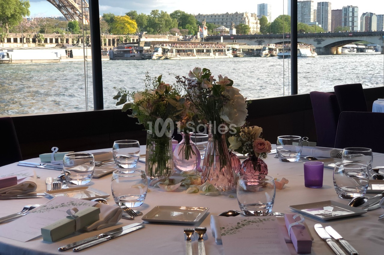 Table élégamment dressée avec des fleurs au centre, vue sur la Seine, la tour Eiffel et des bâtiments en arrière-plan.