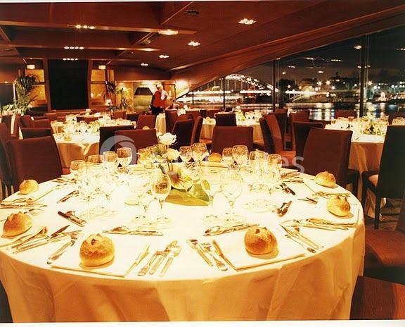 Salle de restaurant élégante avec tables rondes dressées, nappes blanches, verres et couverts, vue nocturne sur l'extérieur.