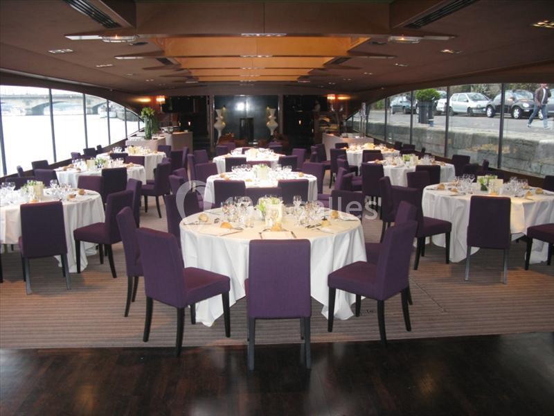 Salle de réception élégante avec tables rondes dressées, nappes blanches et chaises violettes, vue sur l'extérieur.
