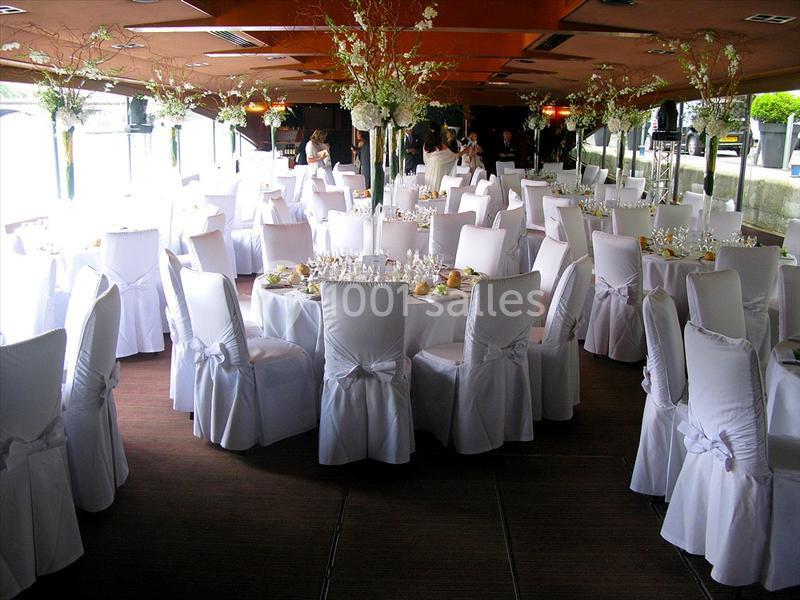Salle de réception décorée avec des tables rondes, nappes blanches et chaises couvertes, prête pour un événement.