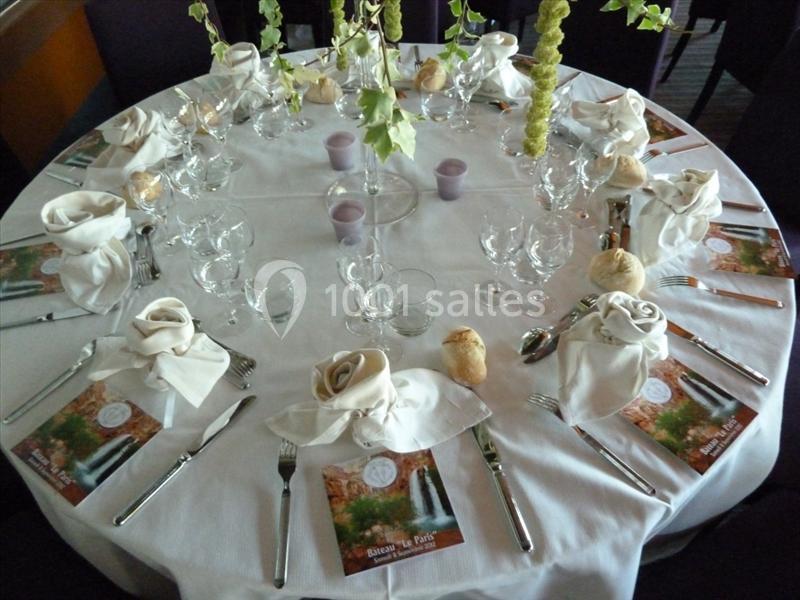 Table ronde dressée avec une nappe blanche, serviettes pliées en forme de rose, verres, couverts et petits pains.