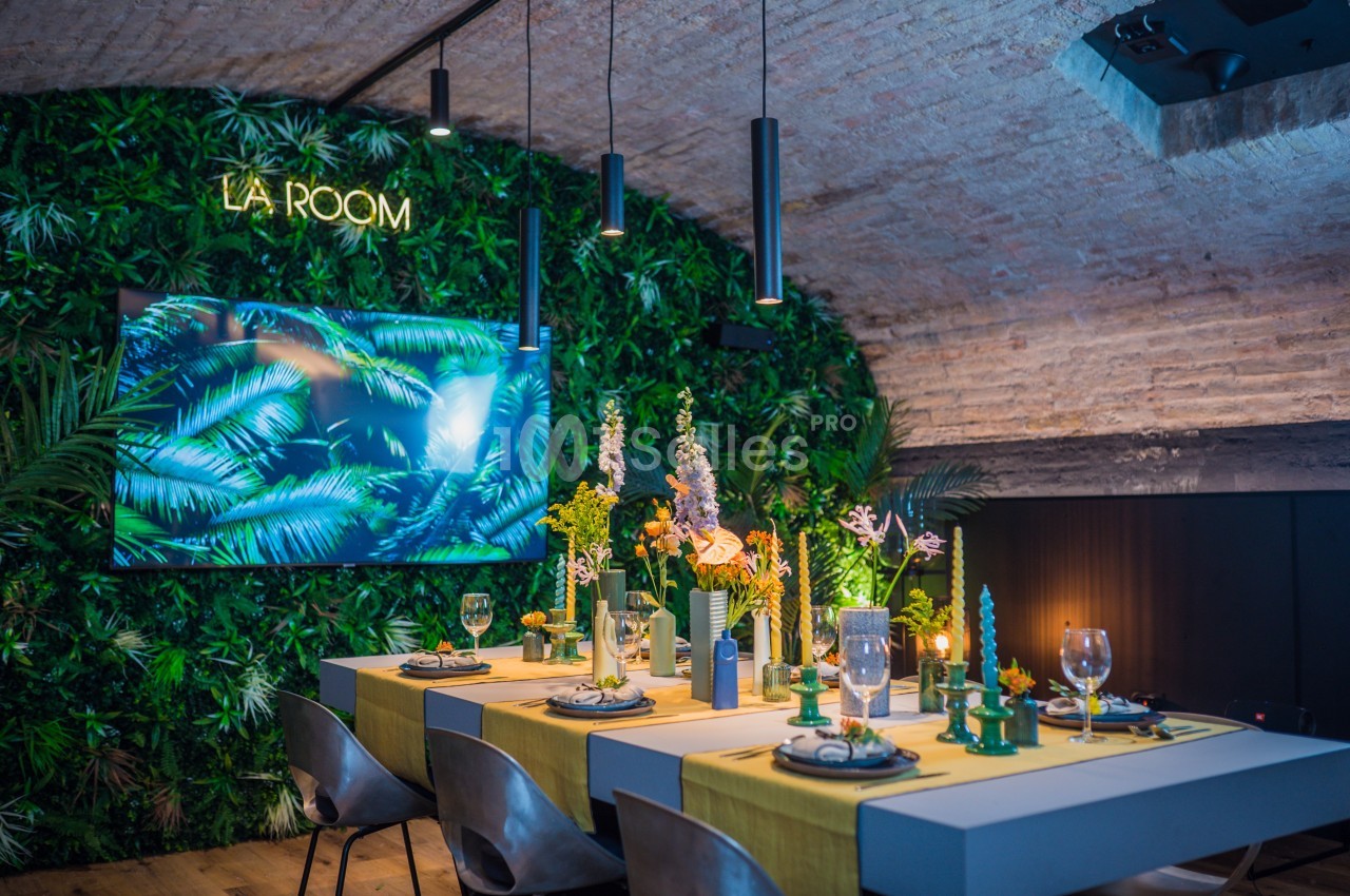 Salle voûtée décorée de plantes, avec une table dressée pour un repas et un écran affichant des feuilles tropicales.