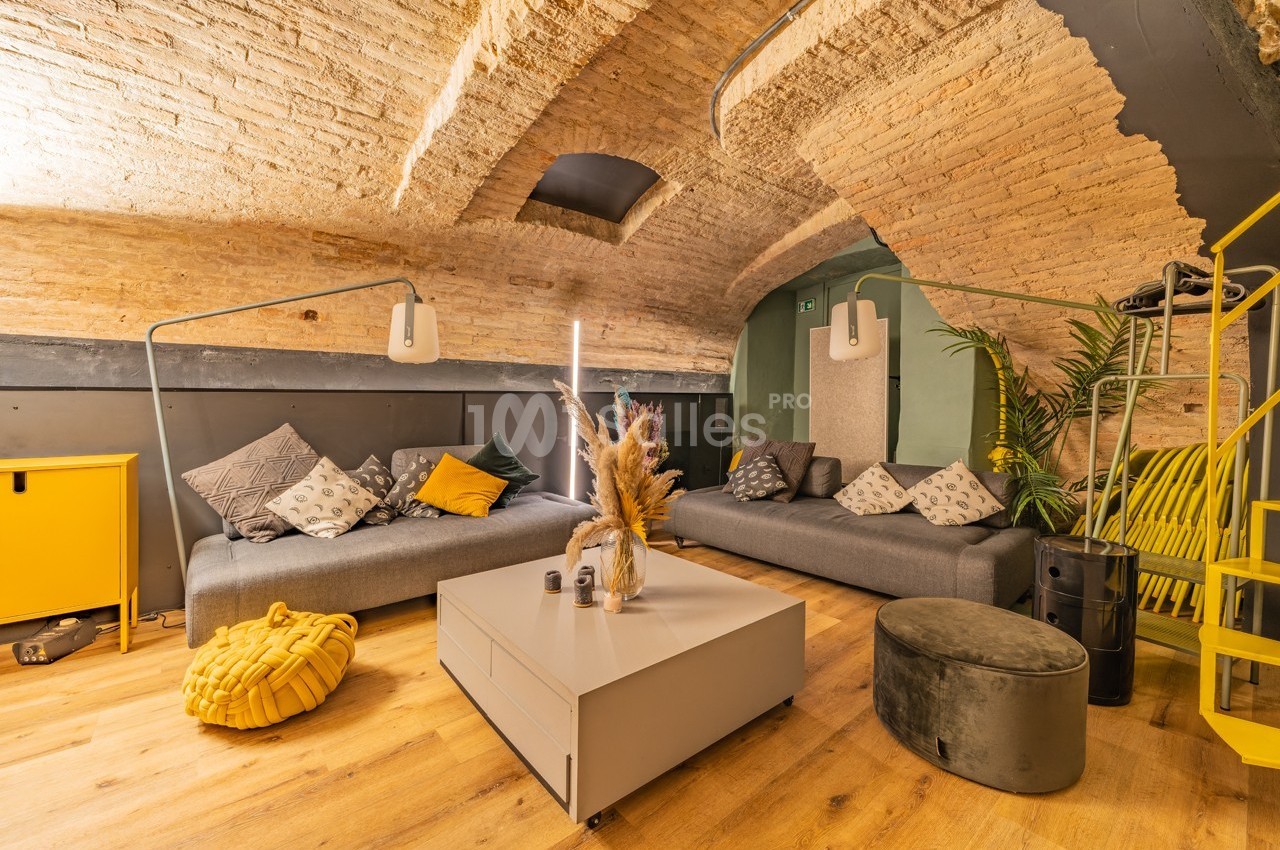 Salon voûté avec murs en briques, canapés gris, table basse moderne et décoration chaleureuse dans des tons jaunes et gris.