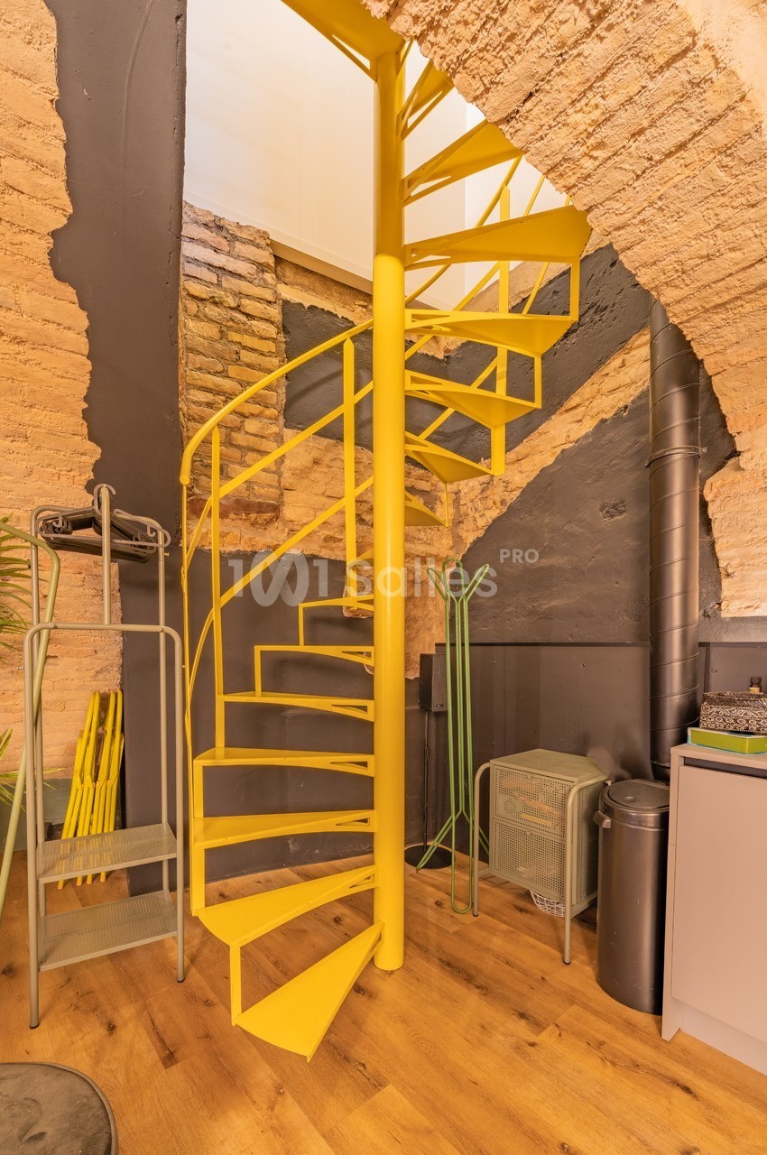 Escalier en colimaçon jaune dans un intérieur avec murs en briques apparentes et parquet en bois clair.