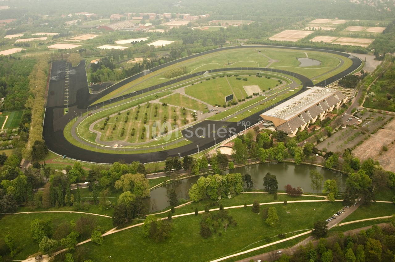 Vue aérienne d'un circuit automobile entouré de verdure, avec des bâtiments et un plan d'eau à proximité.