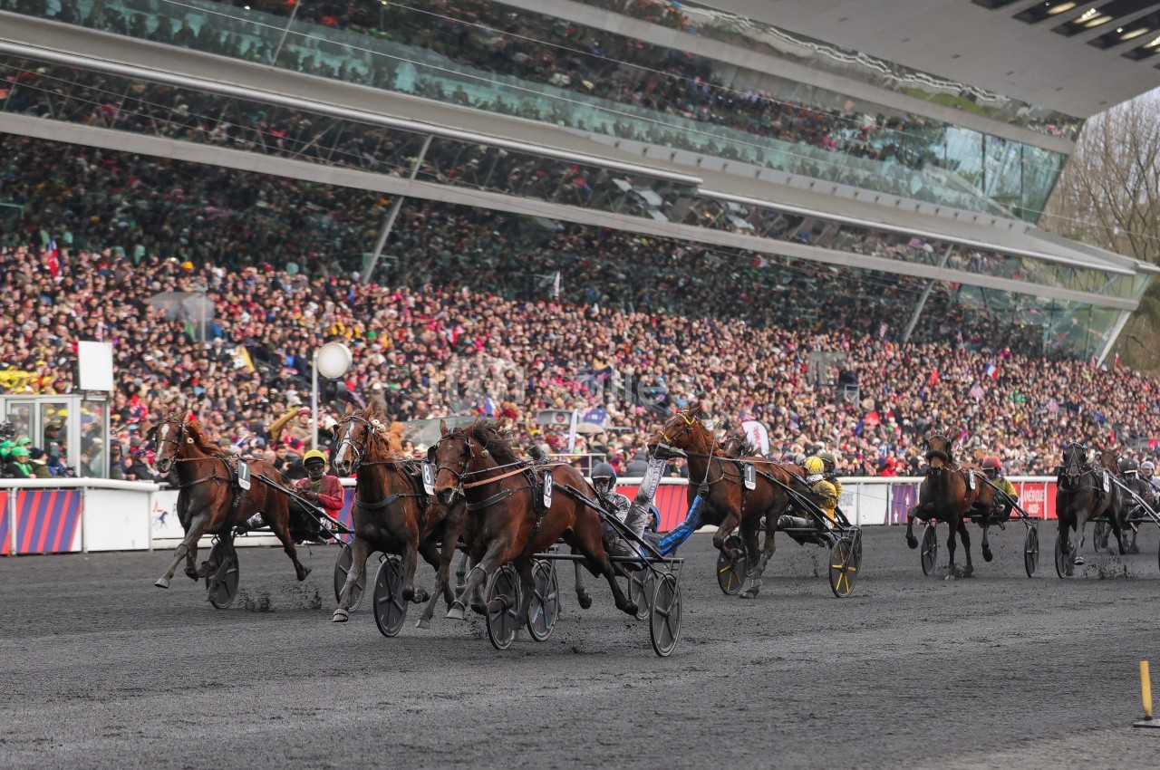 Course de trot attelé sur une piste devant des tribunes pleines de spectateurs.