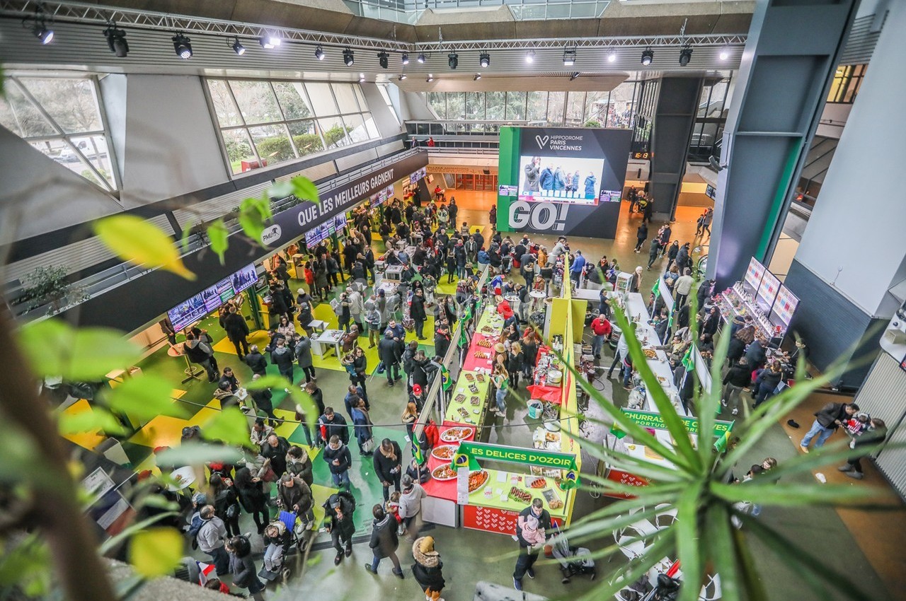 Vue aérienne d'un salon animé avec des stands colorés, des visiteurs et une grande bannière affichant ’GO!’.