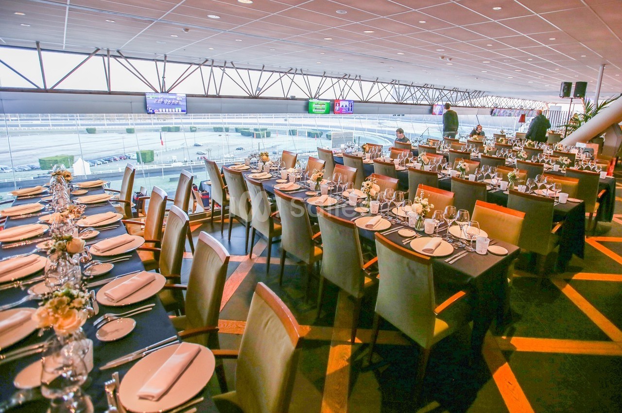 Salle de réception avec de longues tables dressées, vue sur un espace extérieur à travers de grandes baies vitrées.