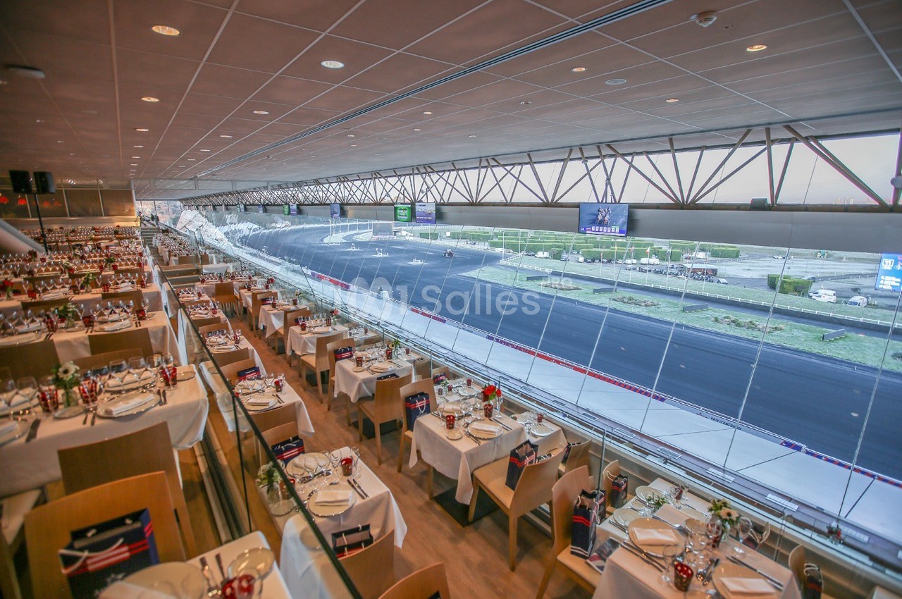 Salle de restaurant avec vue panoramique sur un circuit automobile, tables dressées et baies vitrées.