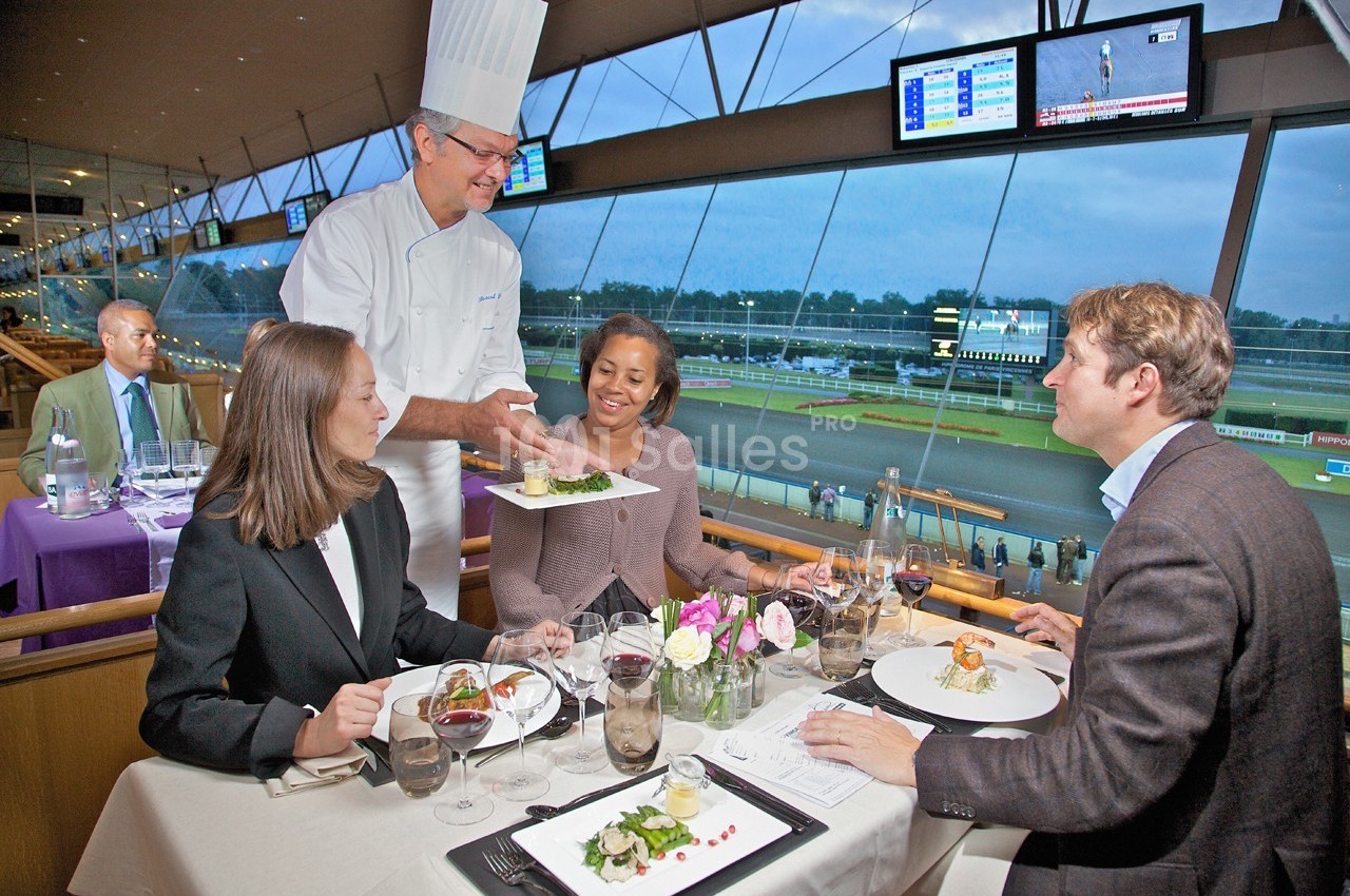 Un chef sert des plats à trois convives attablés dans un restaurant avec vue sur un hippodrome.