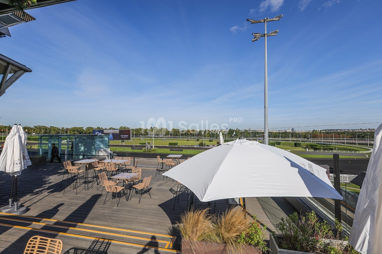 Terrasse en bois avec tables et parasols, vue sur un champ de course sous un ciel bleu dégagé.
