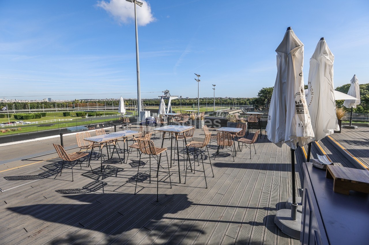 Terrasse extérieure avec tables et chaises en métal, parasols fermés, vue sur un espace vert et ciel dégagé.