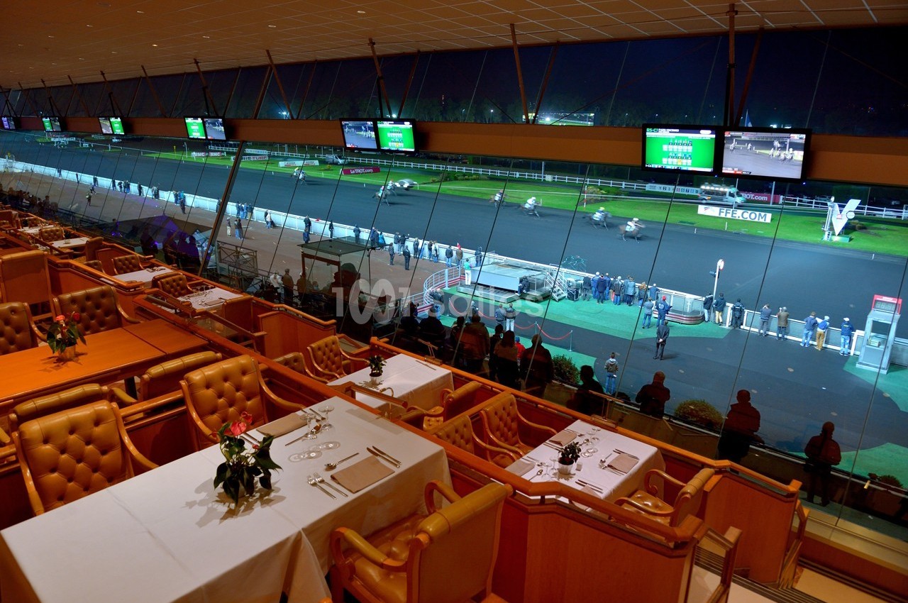 Vue depuis une salle de restaurant surplombant une piste de course hippique éclairée en soirée.