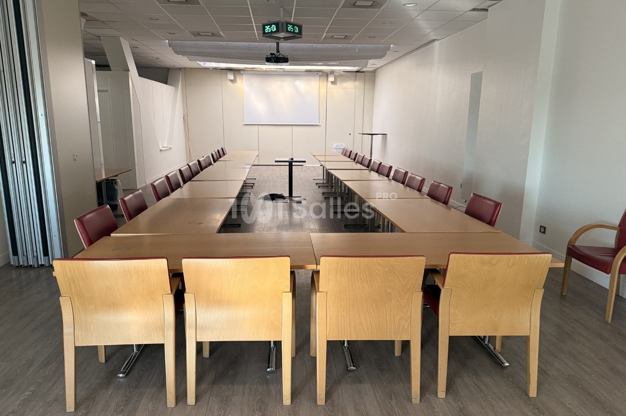 Salle de réunion lumineuse avec tables disposées en U, chaises en bois et écran de projection au fond.