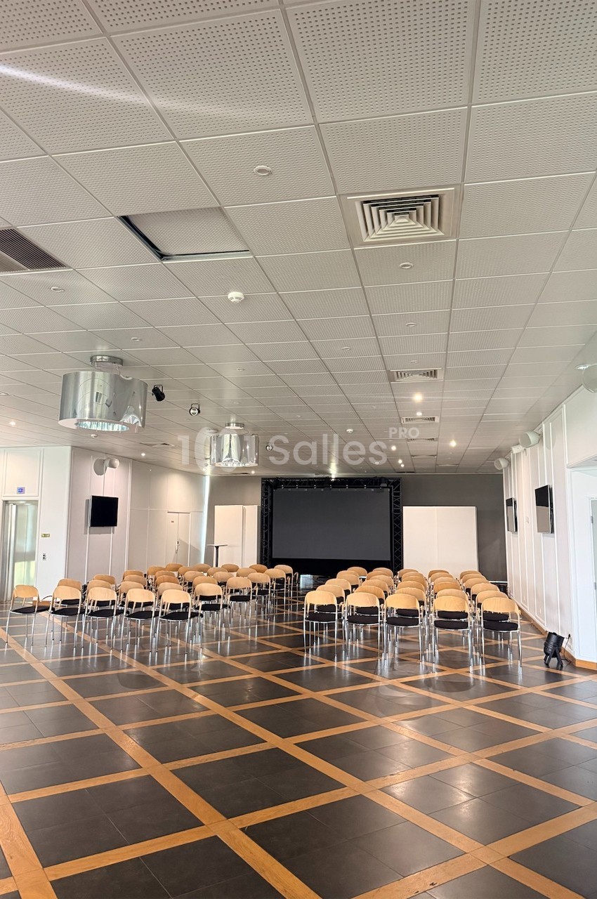Salle de conférence vide avec des rangées de chaises face à un écran noir, éclairée par des lumières au plafond.