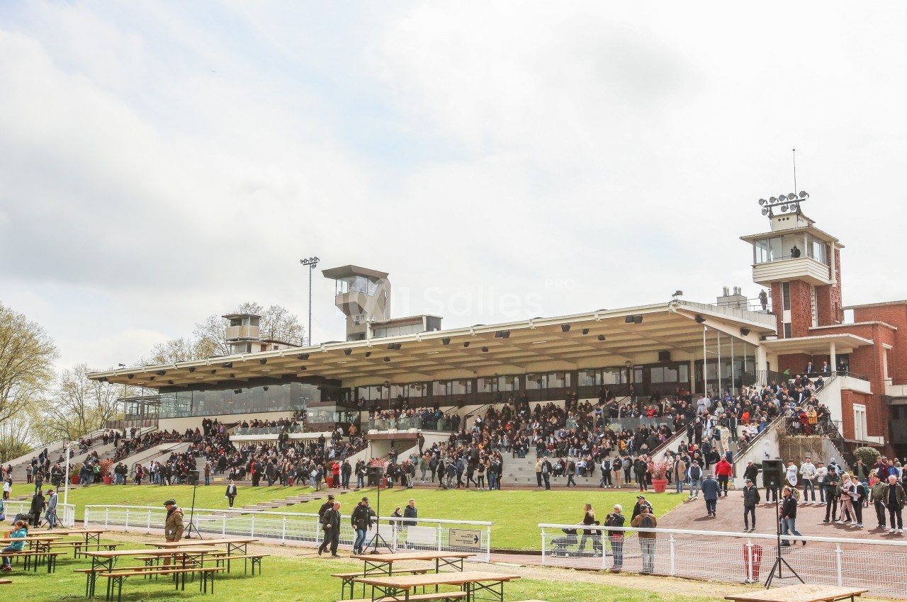 Tribune d'un hippodrome avec des spectateurs assis et debout, entourée d'espaces verts et de tables en plein air.