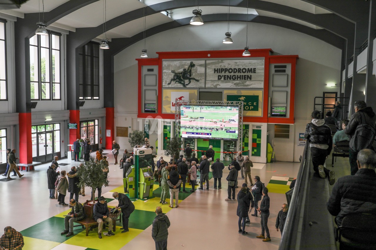 Vue intérieure d'un hall avec des écrans diffusant des courses hippiques, des spectateurs rassemblés au centre.