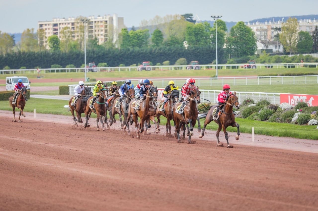 Course de chevaux sur une piste en terre battue, avec des jockeys en tenue colorée et des bâtiments en arrière-plan.