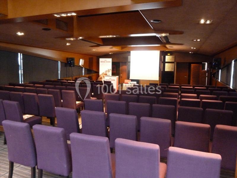 Salle de conférence vide avec des rangées de chaises violettes et un écran de projection allumé à l'avant.