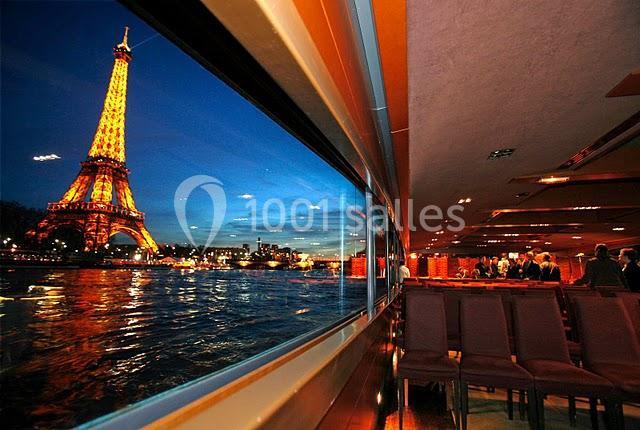 Vue de la tour Eiffel illuminée depuis l'intérieur d'un bateau sur la Seine, avec des reflets sur l'eau.
