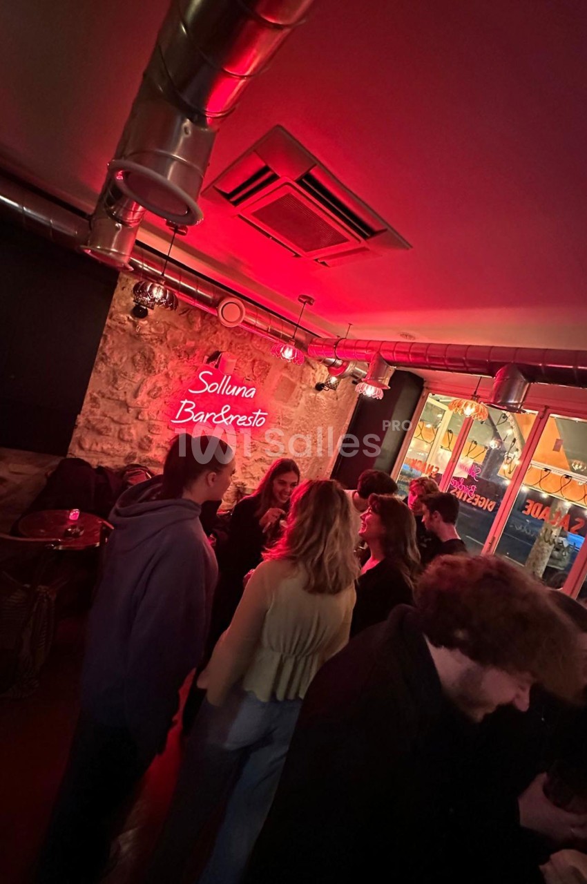 Groupe de personnes discutant dans un bar à l'éclairage rouge avec un mur en pierre et une enseigne lumineuse.