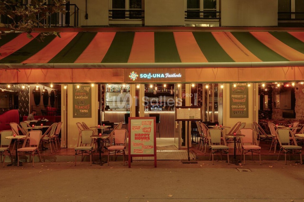 Terrasse d'un restaurant avec chaises en osier et tables dressées, éclairée par des guirlandes lumineuses en soirée.