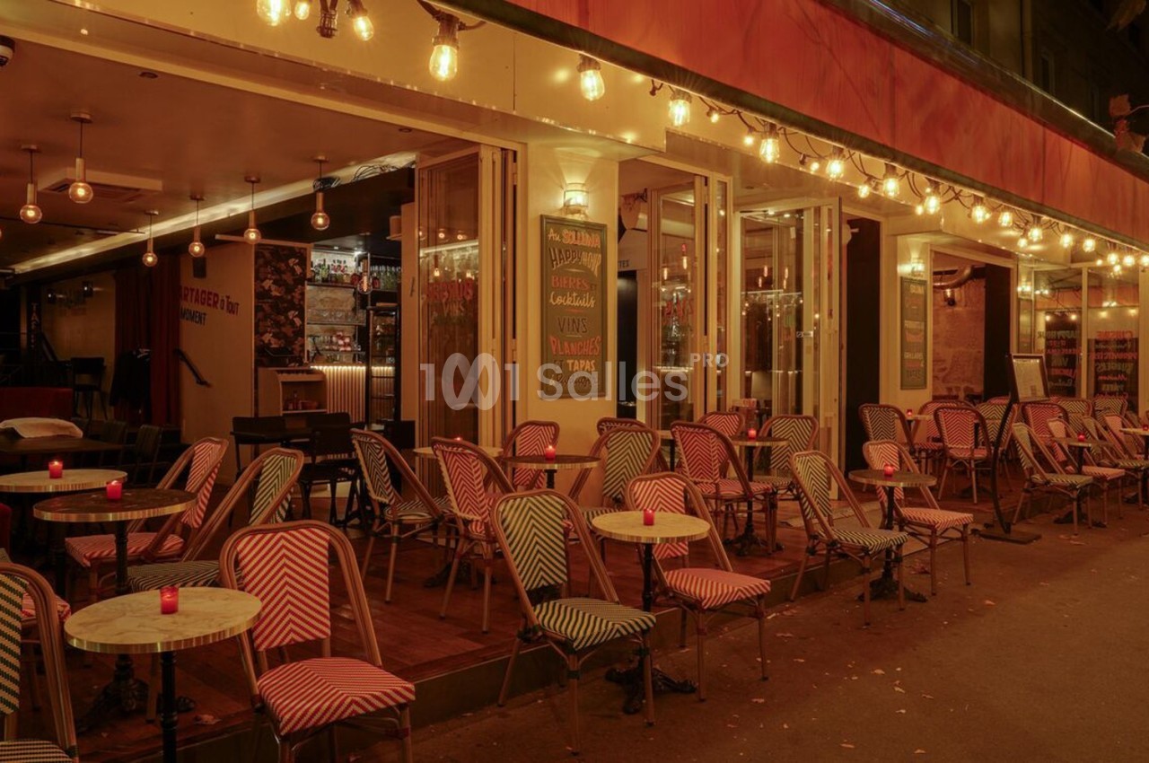 Terrasse d’un café-restaurant éclairée par des guirlandes lumineuses, avec des chaises et tables vides en soirée.