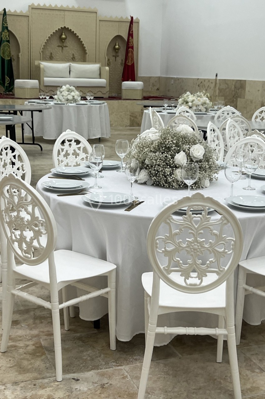 Salle décorée pour un événement avec des tables rondes dressées, chaises blanches sculptées et fleurs blanches au centre.