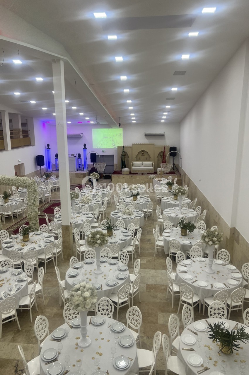 Salle de réception décorée avec des tables rondes, nappes blanches, chaises assorties et arrangements floraux.