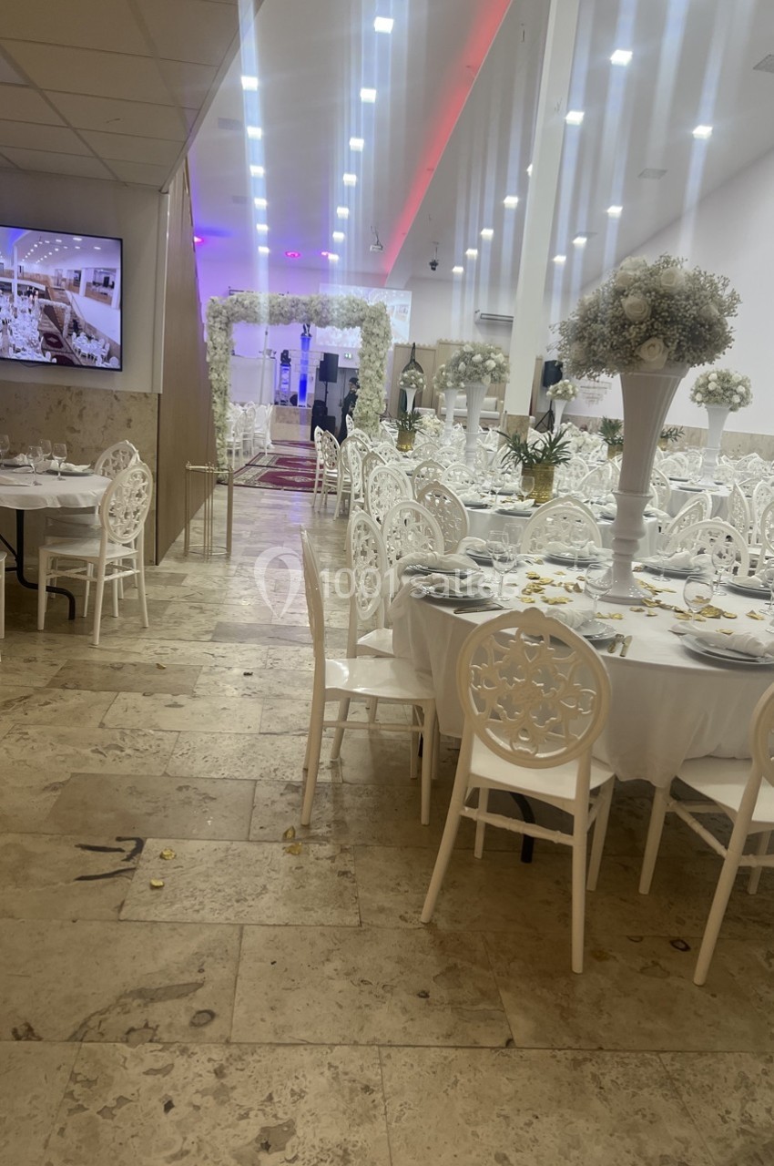 Salle de réception décorée avec des tables rondes, des chaises blanches et des arrangements floraux blancs.
