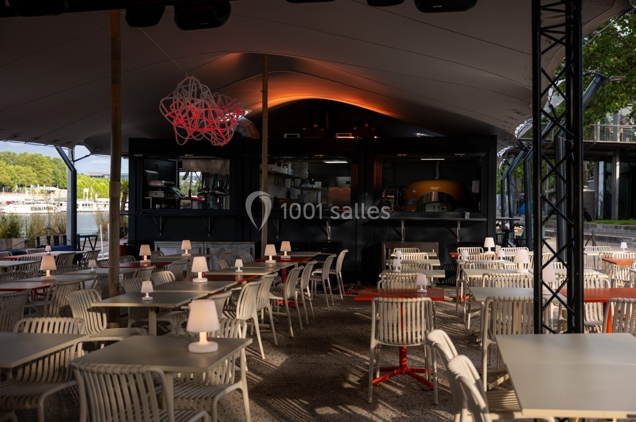 Terrasse couverte d'un restaurant avec des tables et chaises vides, éclairée par des lampes et un four visible en arrière…