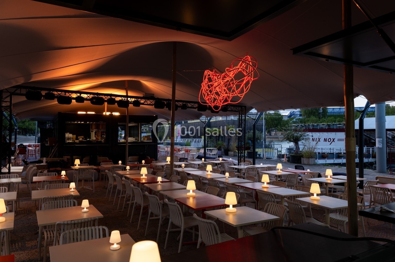 Terrasse couverte avec tables et chaises blanches, éclairée par des lampes et une sculpture lumineuse rouge suspendue.
