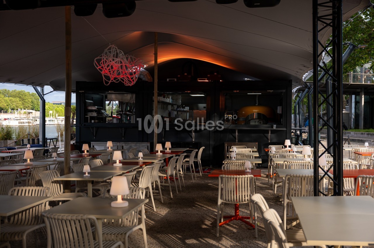 Terrasse couverte d'un restaurant avec des tables et chaises vides, éclairage tamisé et four à pizza visible en arrière-plan.