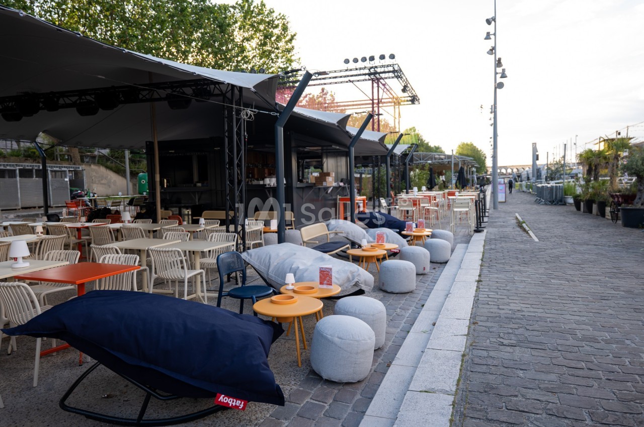Terrasse extérieure avec tables, chaises et poufs colorés, située le long d'une promenade pavée.