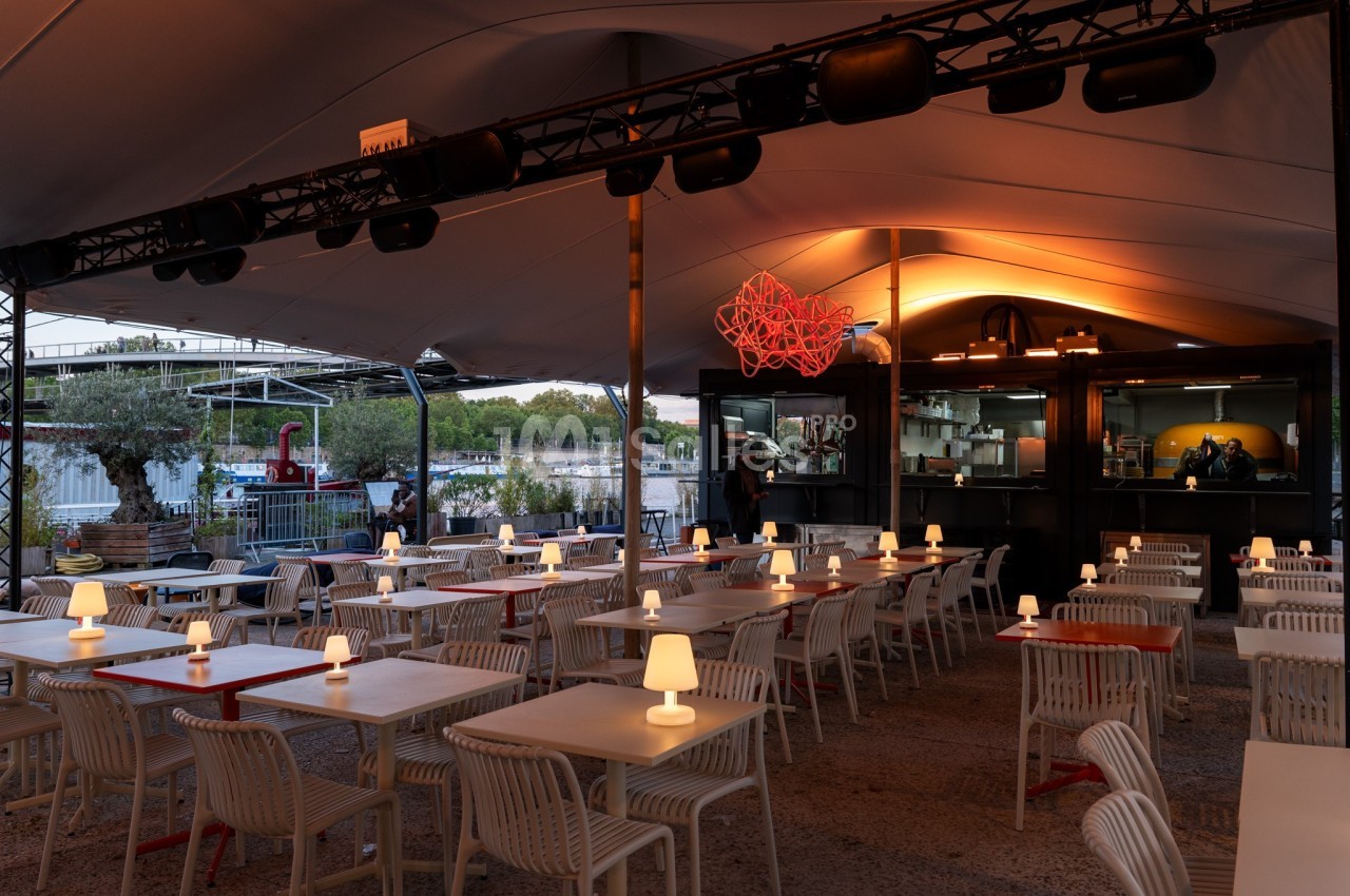 Terrasse de restaurant en soirée avec tables éclairées par des lampes, vue sur un fleuve et cuisine ouverte en arrière-plan.
