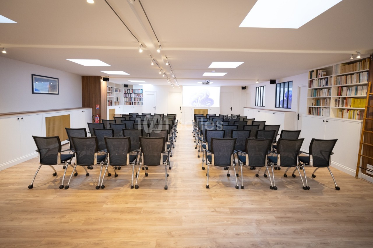 Salle de conférence lumineuse avec des rangées de chaises noires, un écran de projection et des étagères remplies de livres.