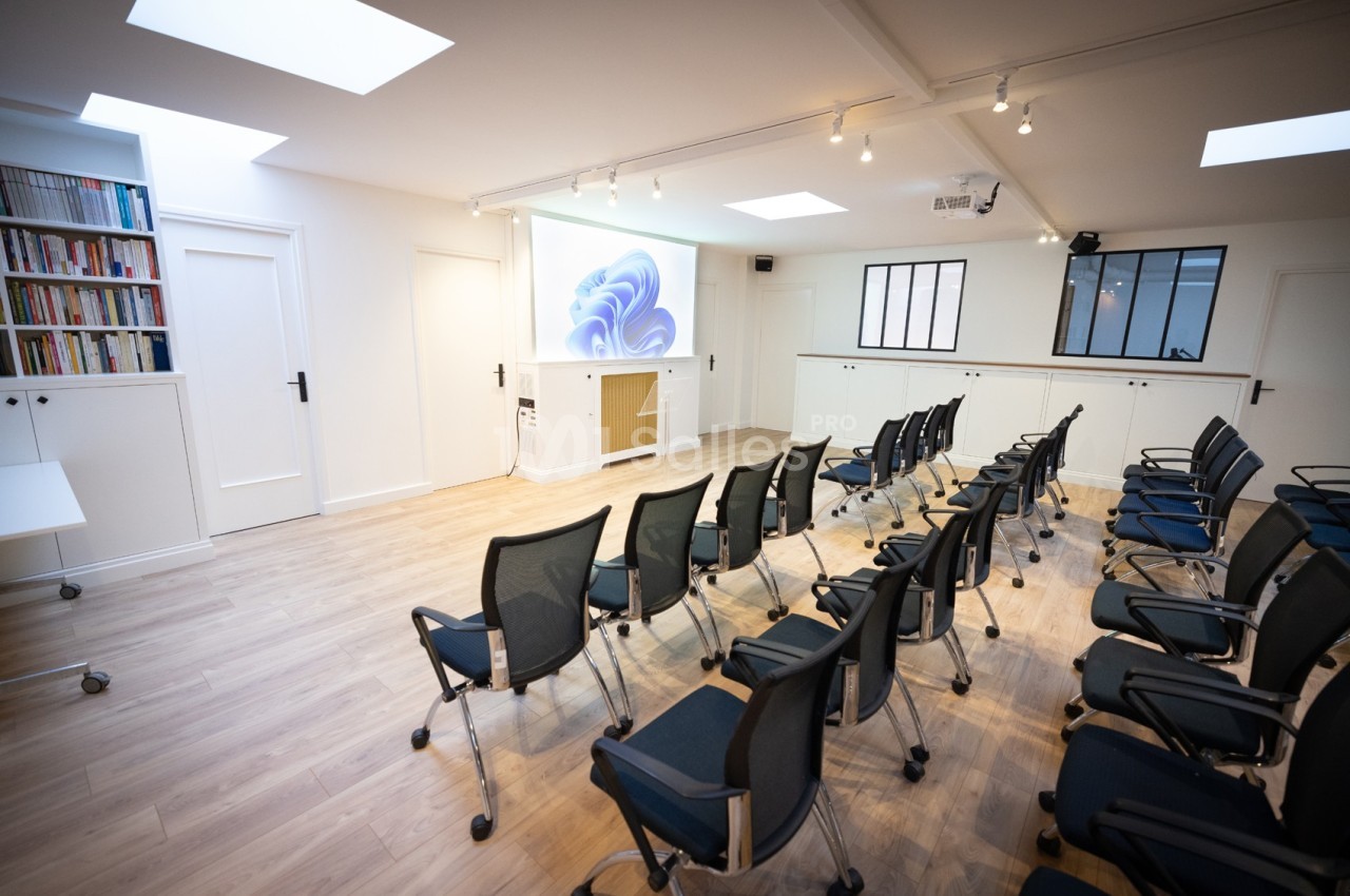 Salle lumineuse avec rangées de chaises noires, écran de projection allumé et étagères remplies de livres.