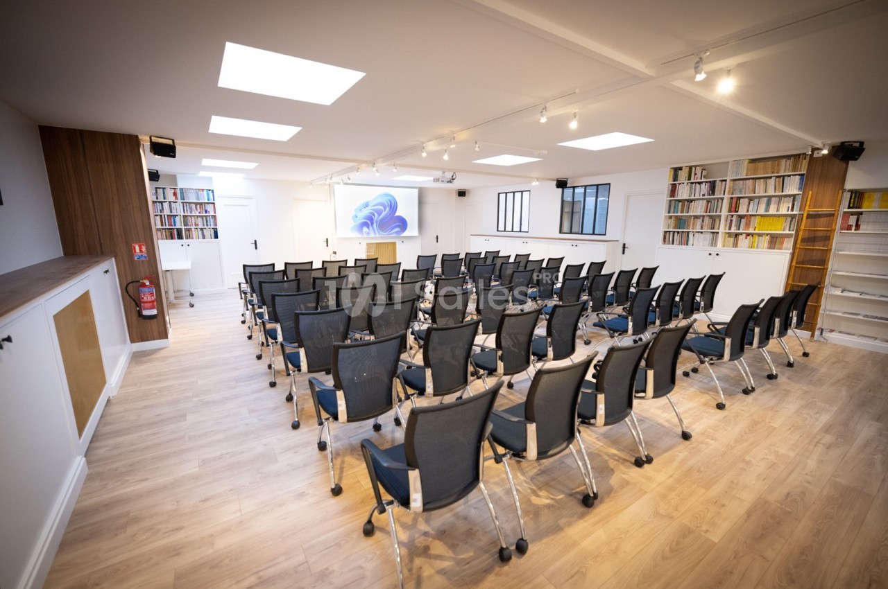 Salle de conférence lumineuse avec rangées de chaises noires, parquet clair et étagères remplies de livres.