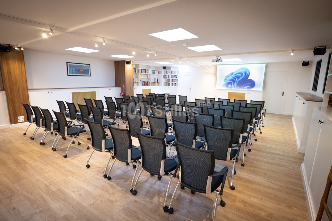 Salle de conférence lumineuse avec des rangées de chaises noires, un écran de projection et des étagères remplies de livres.